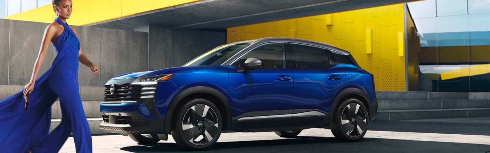 Blue 2025 Nissan Kicks with yellow building in background and lady in blue dress
